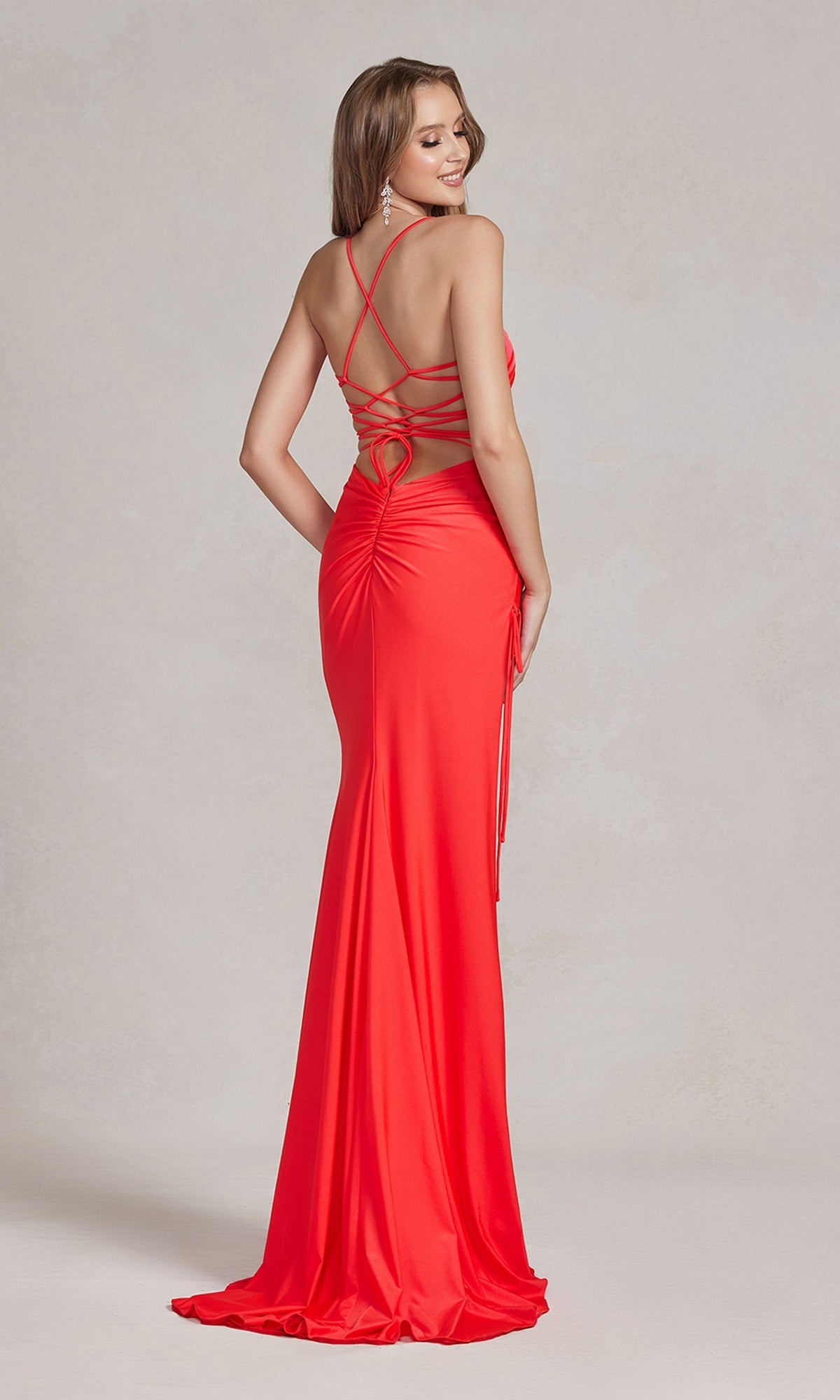 A woman models the Strappy-Back Affordable Long Prom Dress in bright red, featuring crisscross straps and ruched detailing, as she stands and glances over her shoulder.